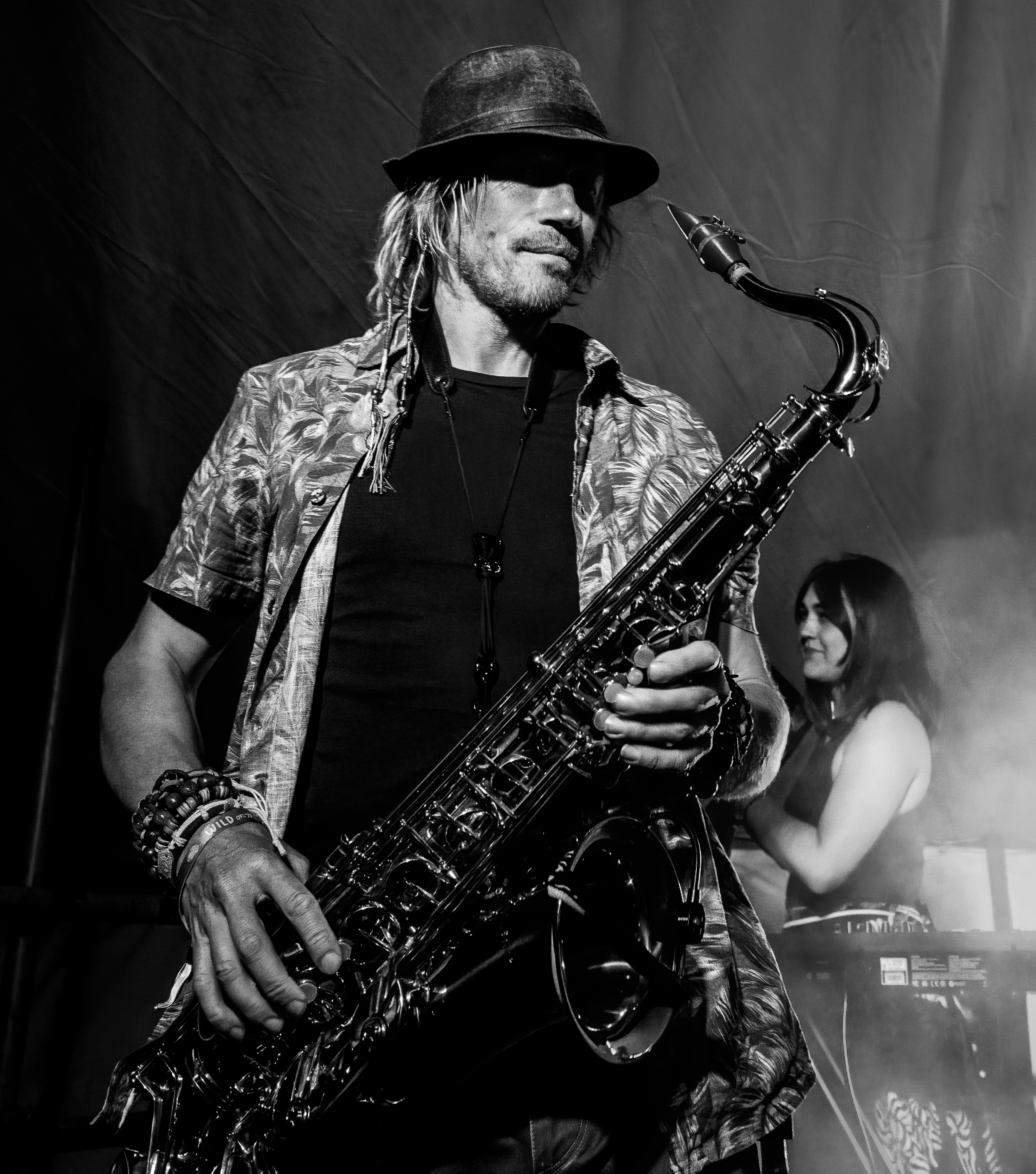 A saxophonist performing on stage, wearing a patterned shirt and a hat, with a female musician playing keyboard in the background. The image is in black and white, capturing a lively concert atmosphere.