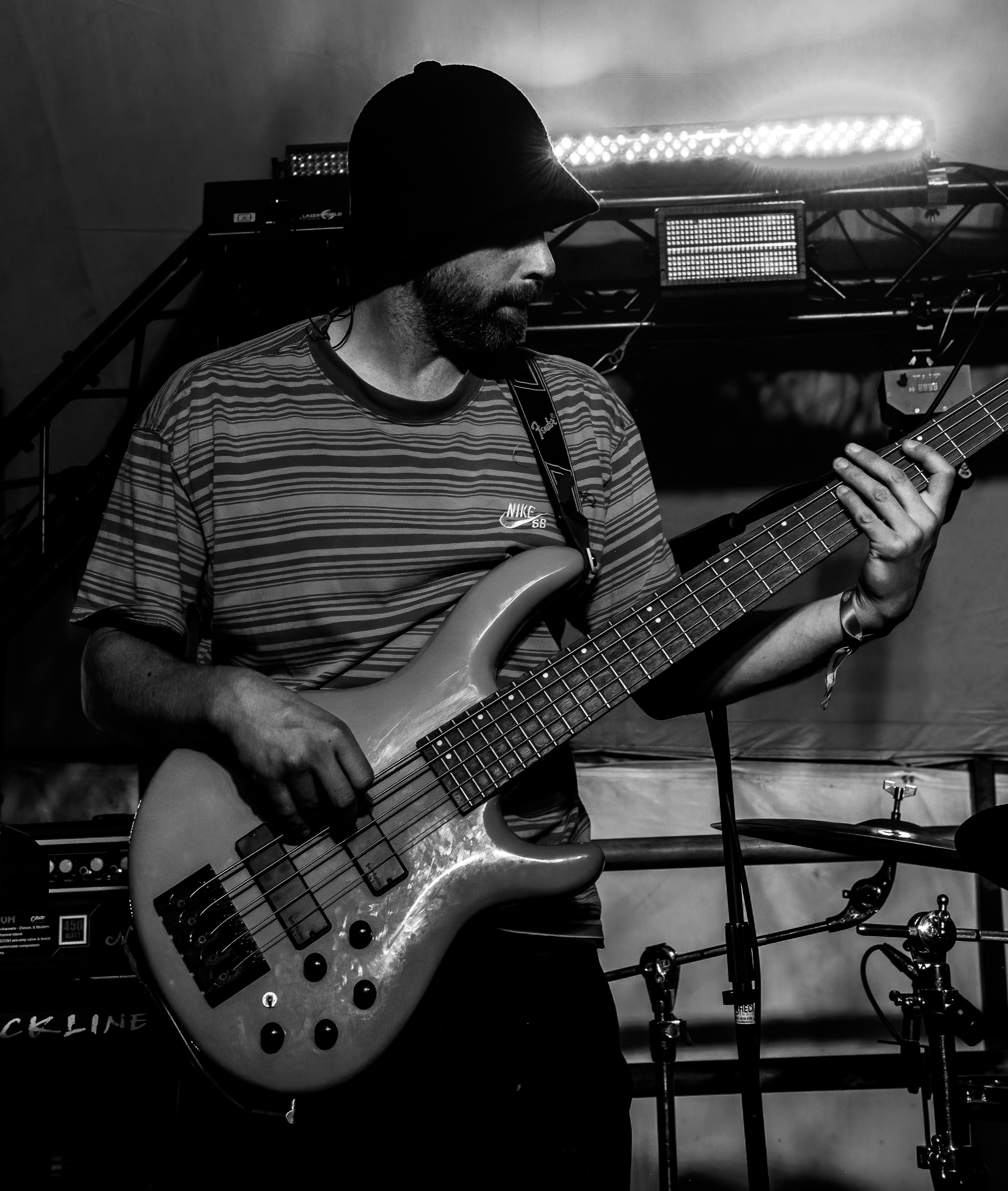 Black and white image of a musician playing a bass guitar on stage, wearing a beanie and a striped shirt.