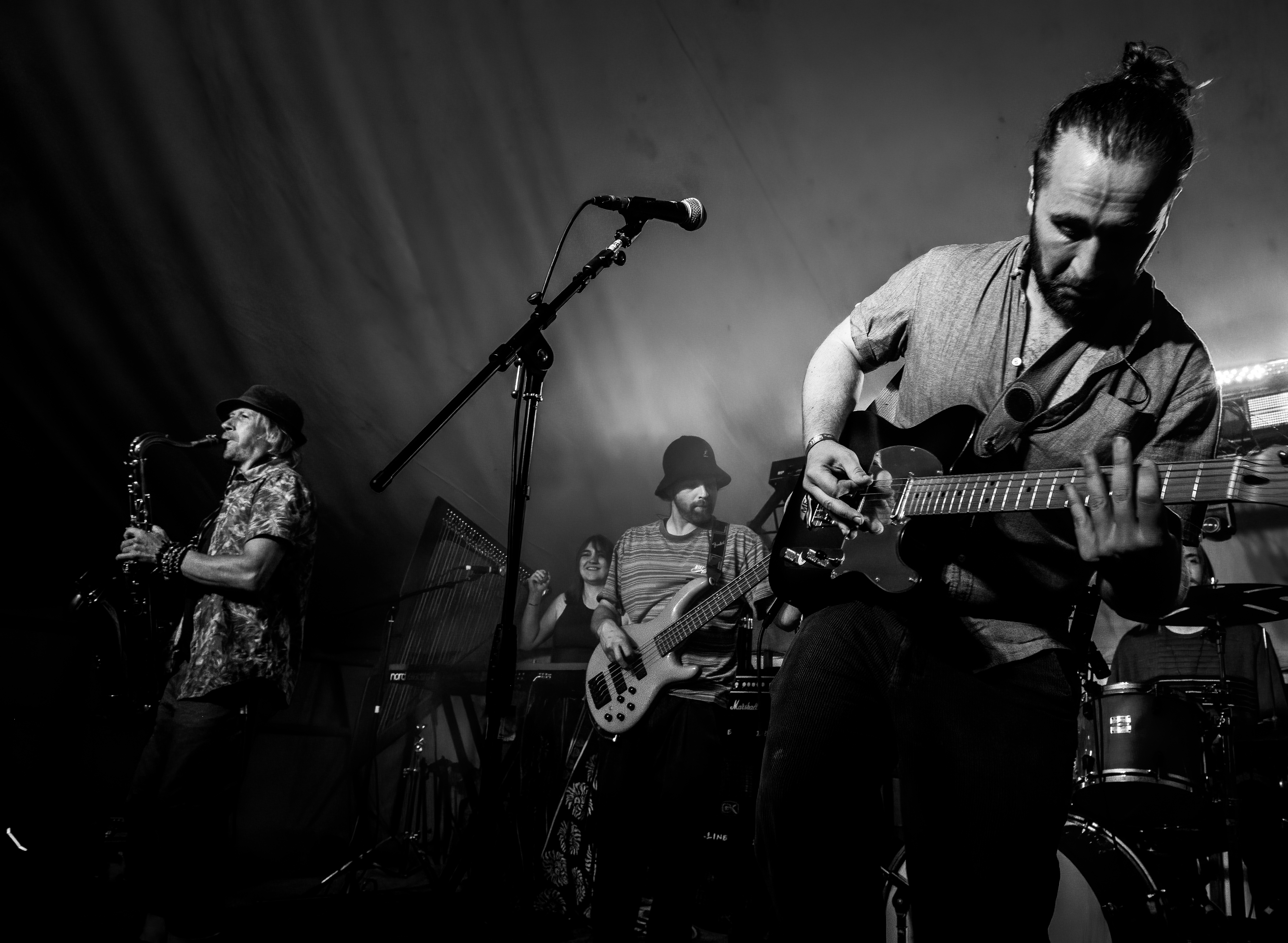 A black and white photo of a live band performing on stage, featuring a saxophonist, a guitarist playing an electric guitar, a bassist, and a keyboardist, with dramatic lighting in a festival tent.