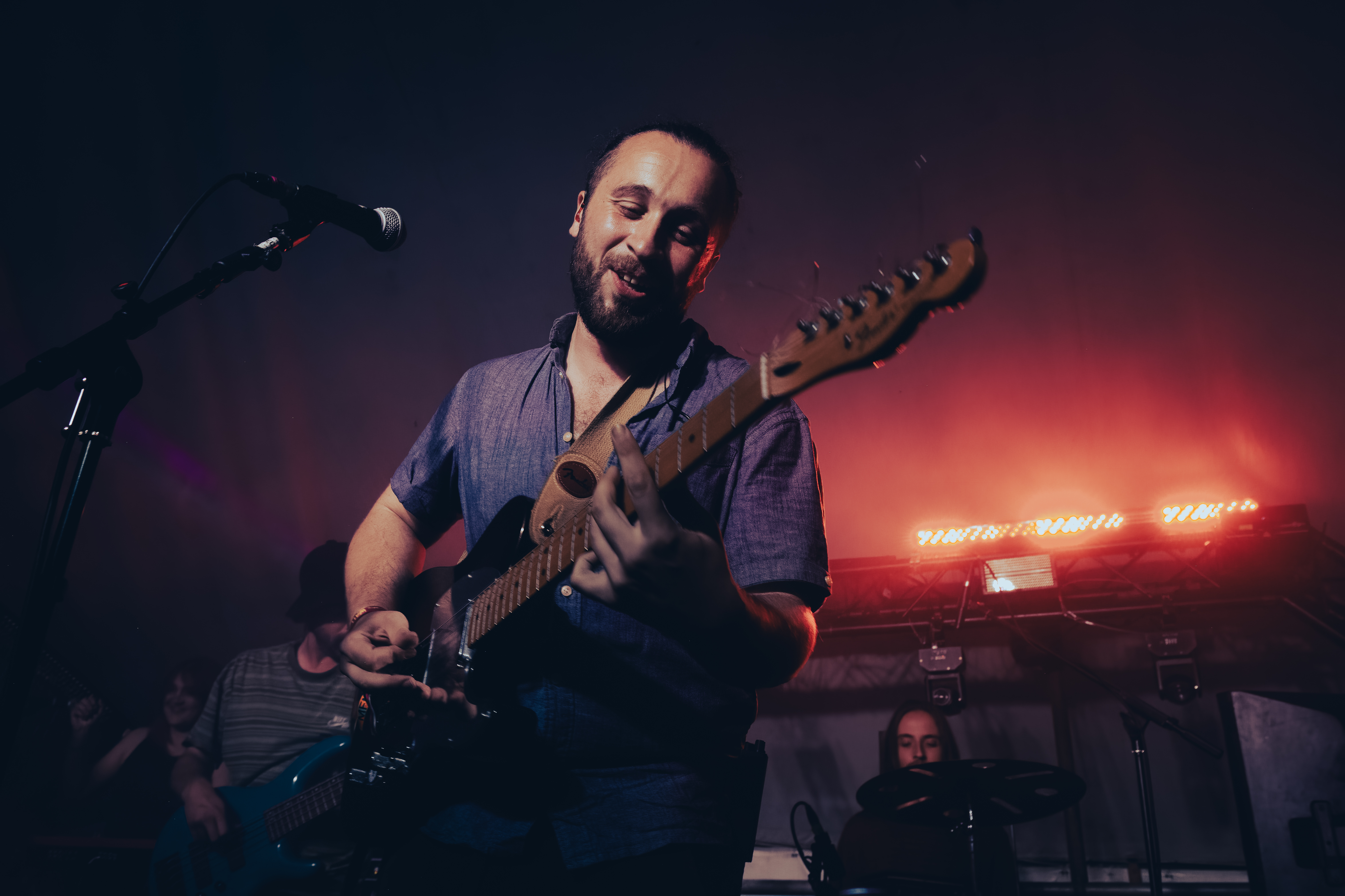 A musician playing an electric guitar on stage, smiling while performing, with colorful stage lights in the background and a drummer visible in the shadows.