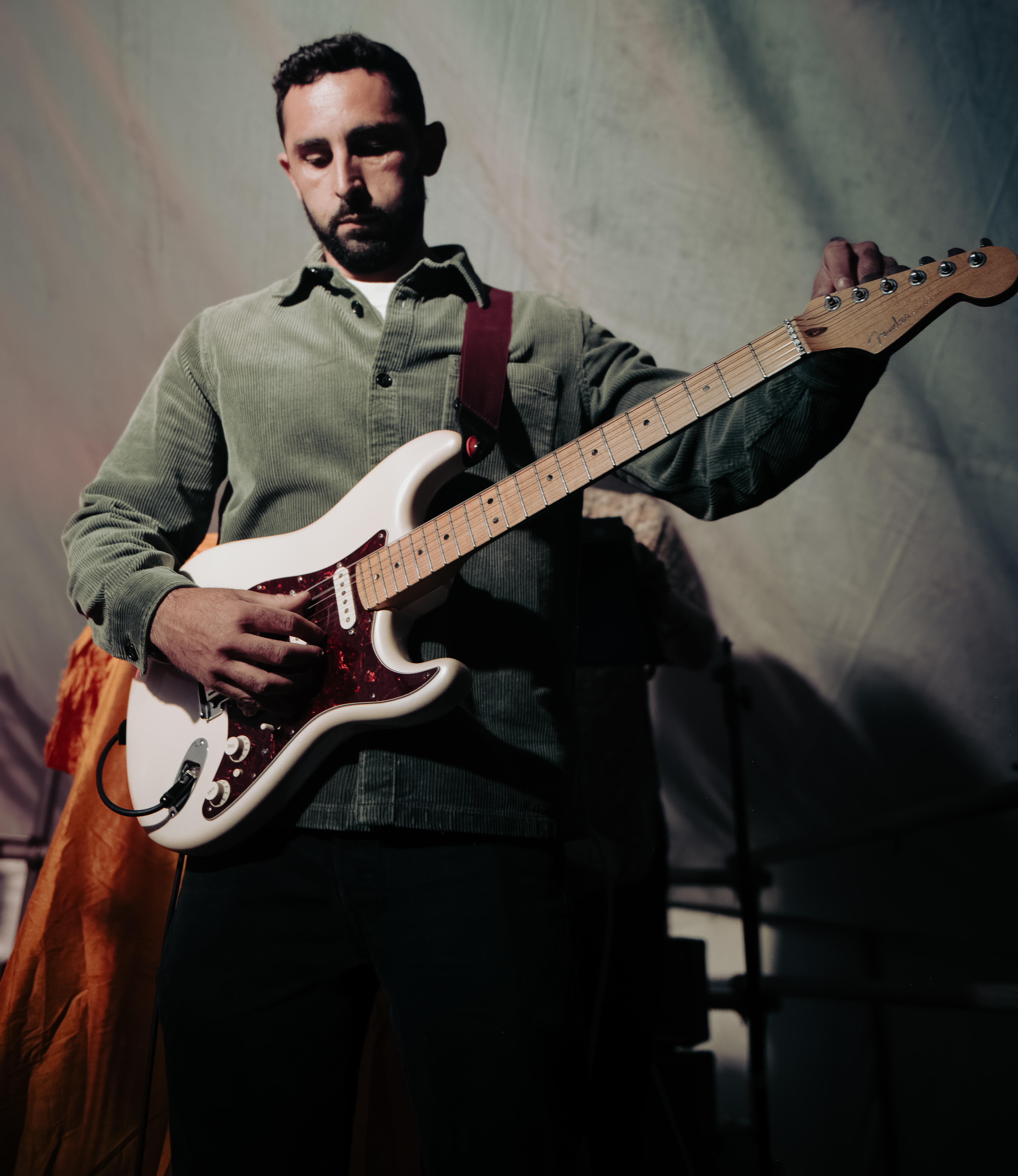 A musician playing a white electric guitar on stage, wearing a green corduroy shirt.