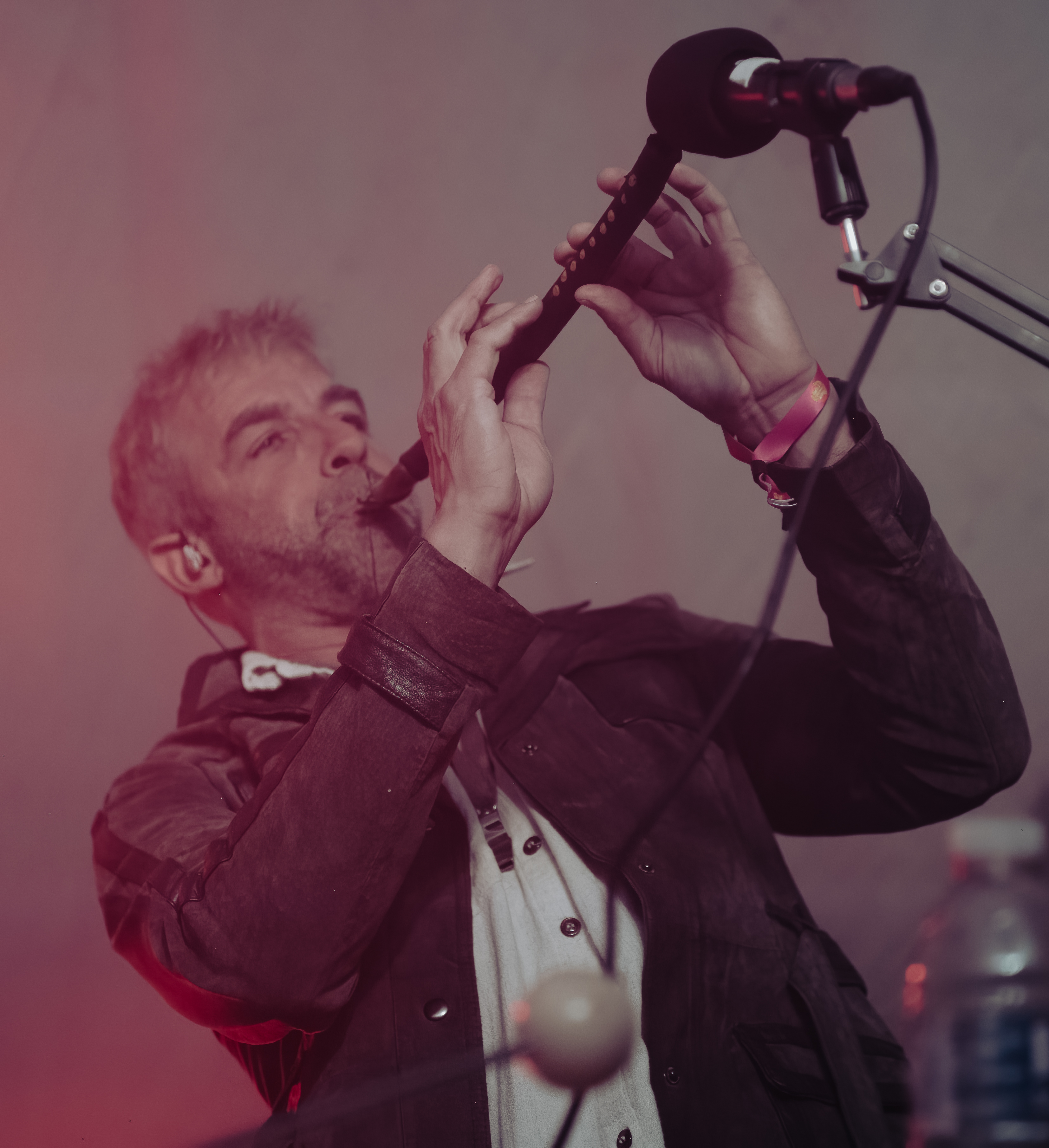 A man playing a flute on stage, wearing a leather jacket and an ear monitor, illuminated by soft stage lights.