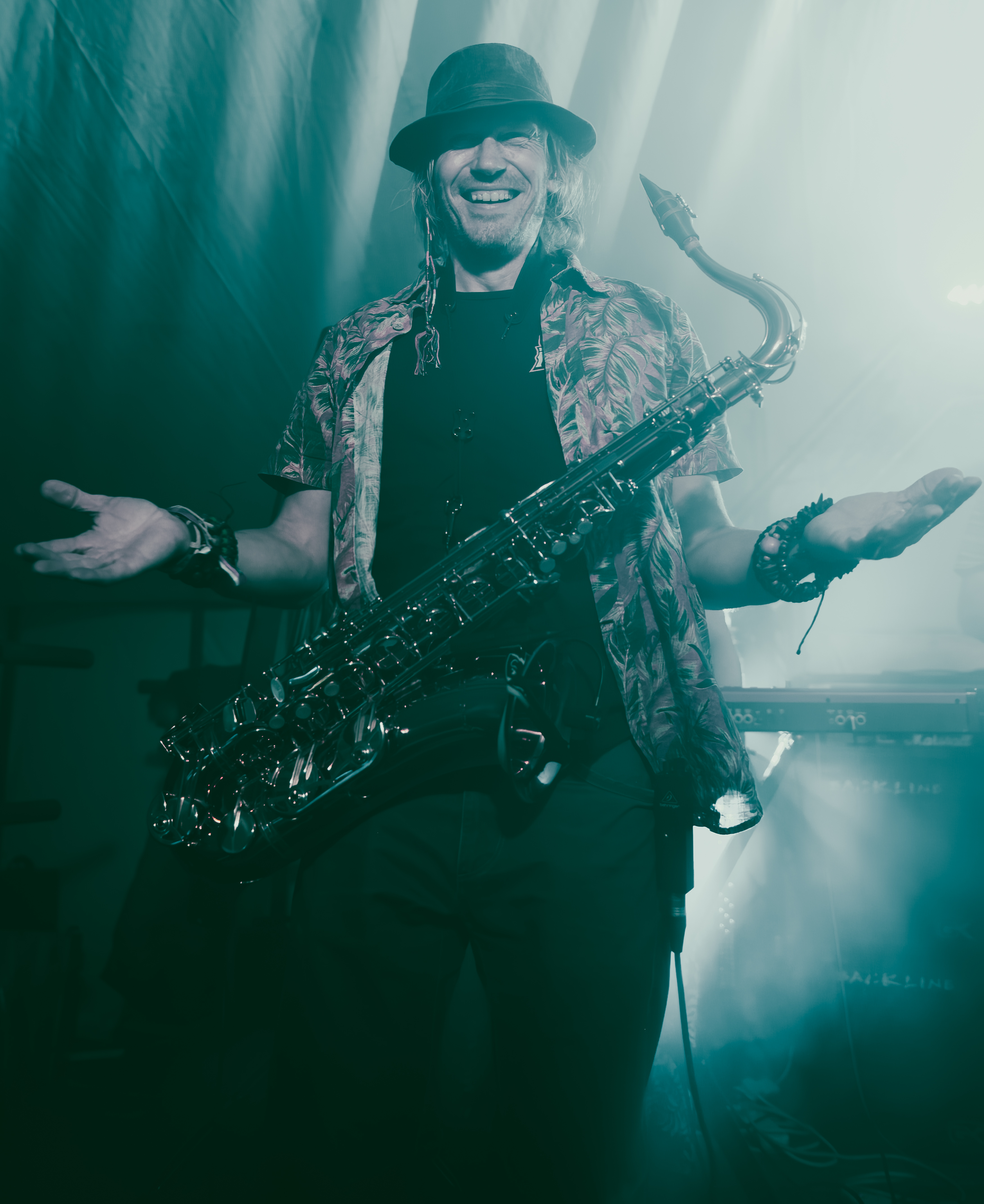 A musician wearing a hat and a patterned shirt holds a saxophone while smiling and gesturing during a performance, with dim lighting and fog in the background.
