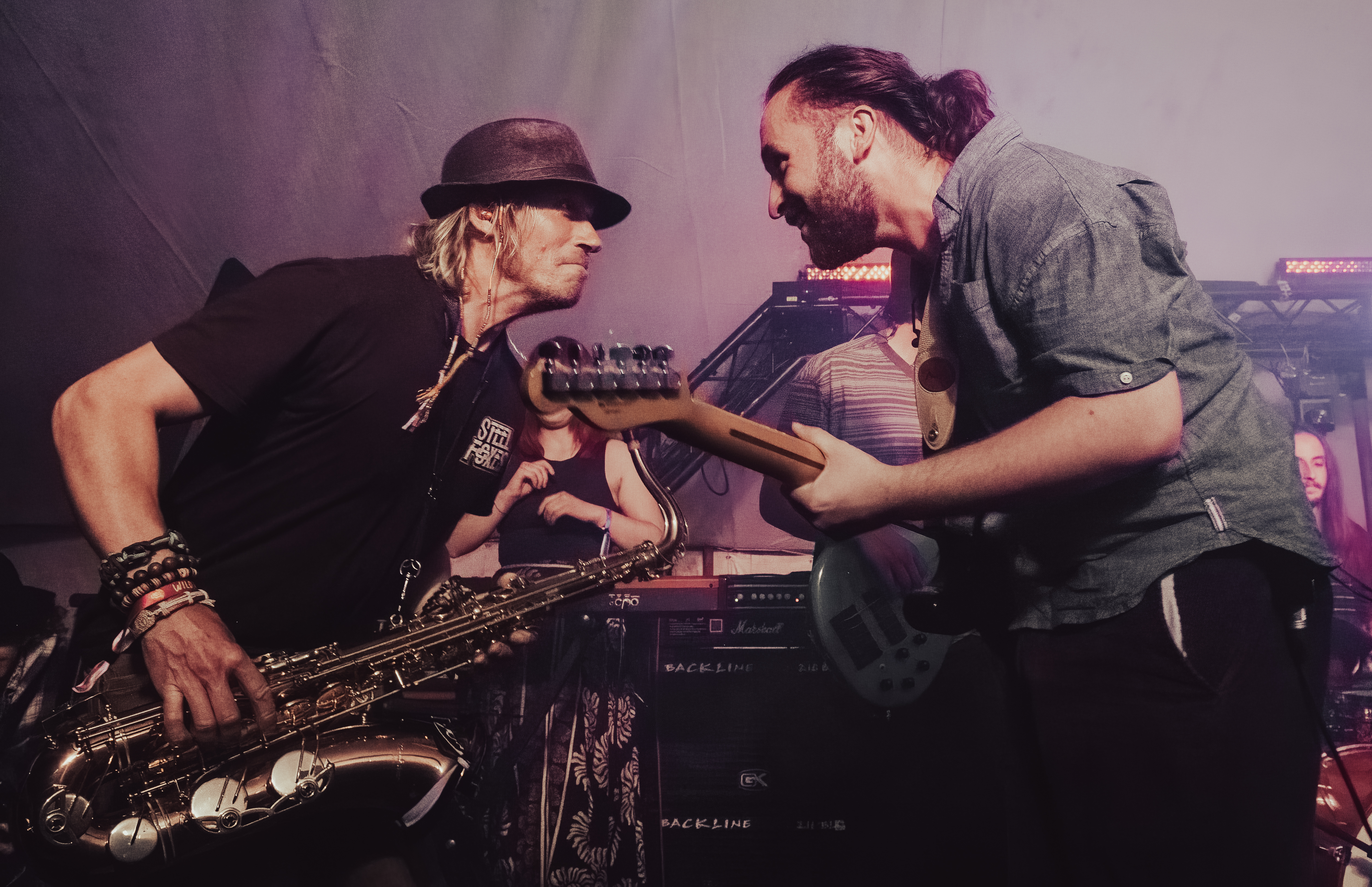 Two musicians performing on stage, one playing the saxophone and the other holding an electric guitar. They display an engaging interaction, with a lively atmosphere emphasized by stage lights in the background.