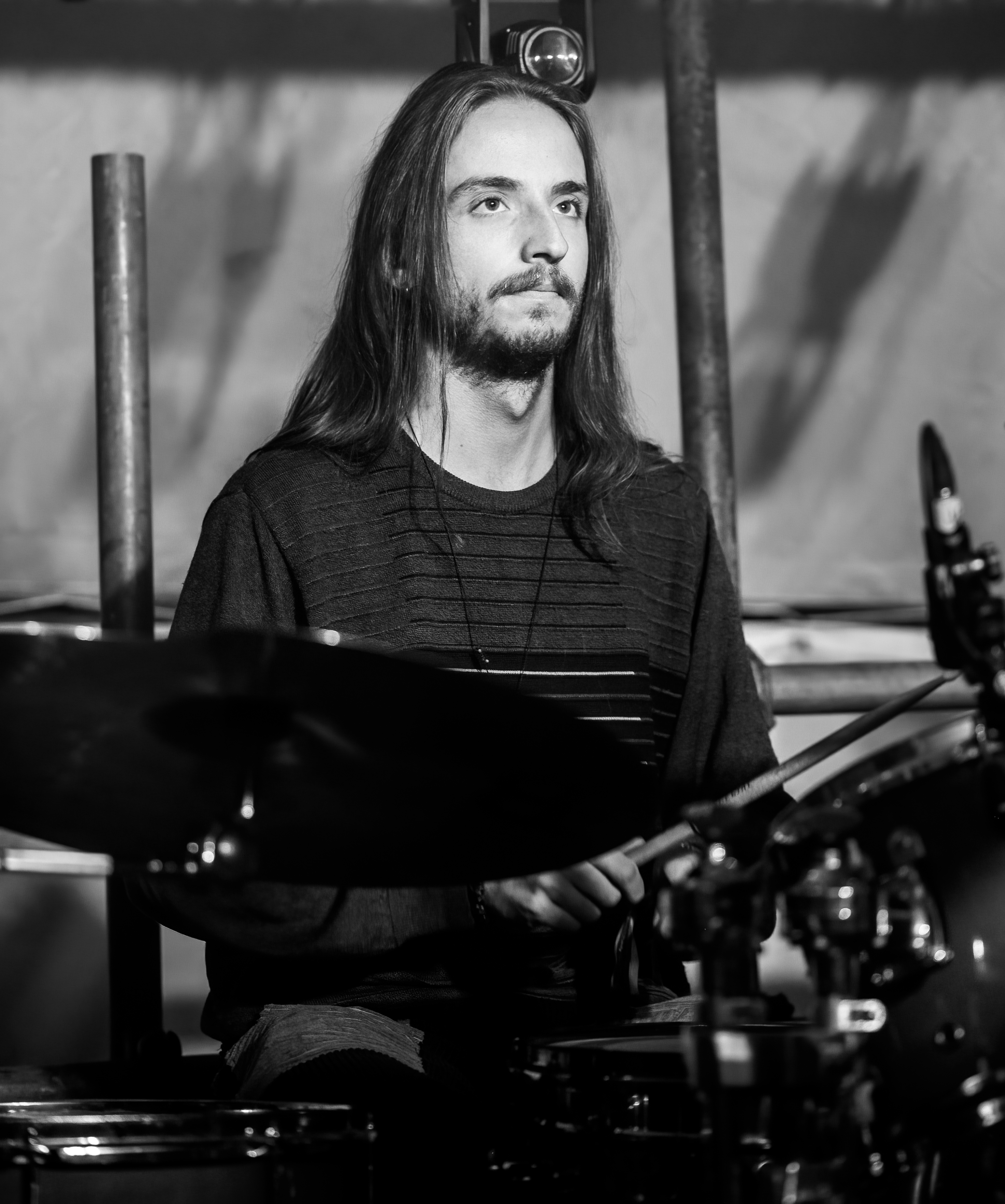 A male drummer with long hair playing drums on stage, focused and serious expression, in black and white.